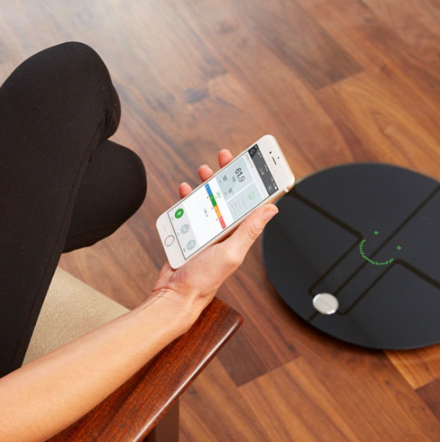A hand holding a phone with an app and the Qardio base smart scale in front.