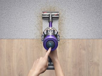 A person vacuums from hardwood to carpet with the Dyson V11 Absolute Pro. They hold the vacuum in their right hand while their left hand points to to the LCD display which reads "Auto."