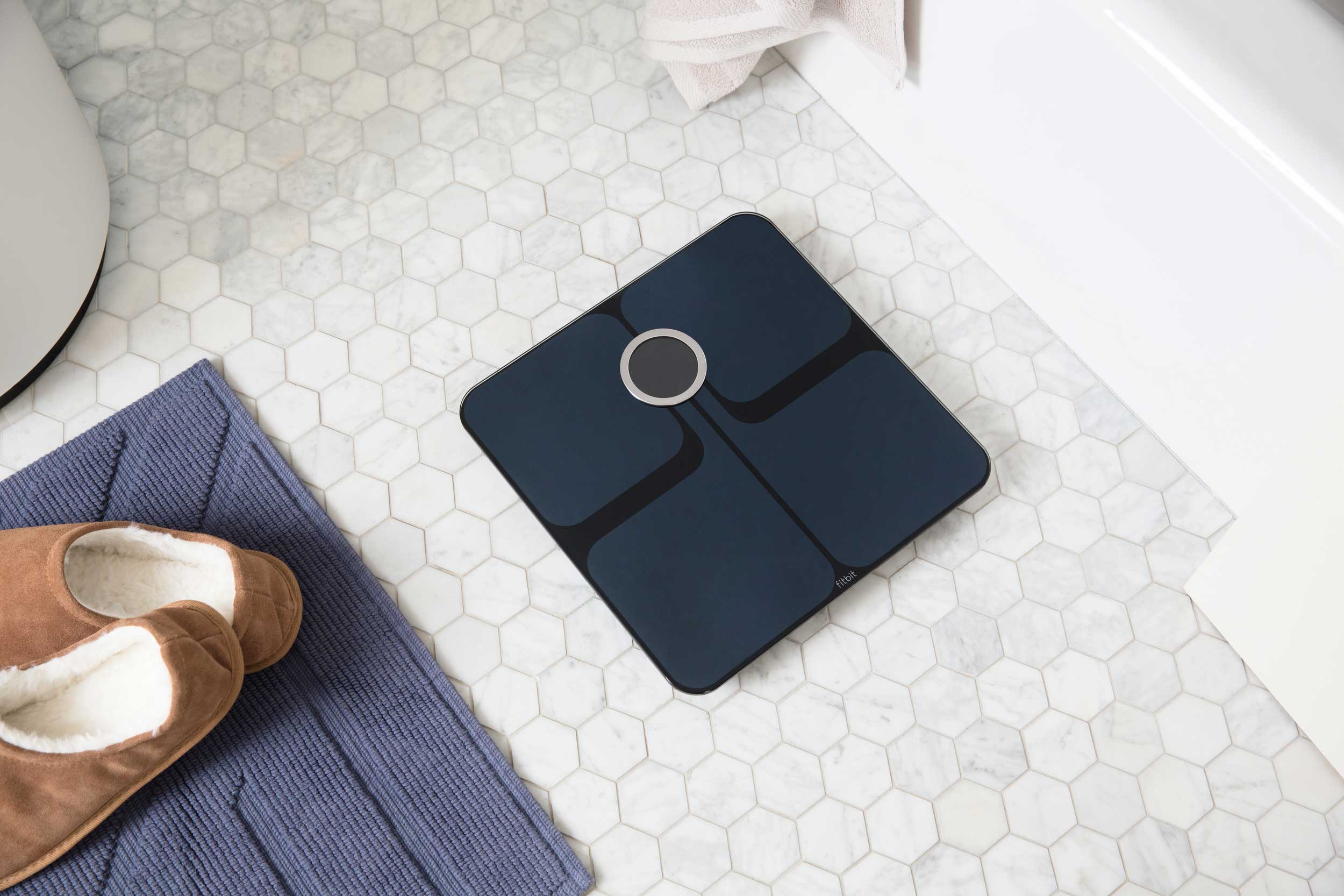 Lifestyle photo of a dark coloured fitbit smart scale on the bathroom floor