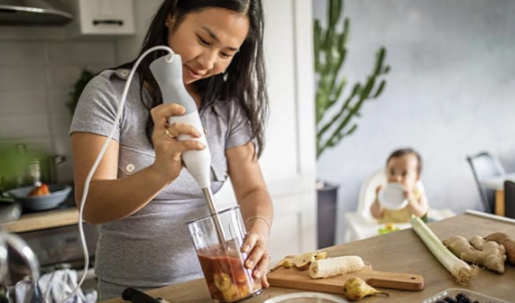 Mom with immersion blender, toddler in the background.