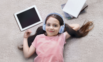 Little girl lying on the floor wearing Kiddesigns kids headphones with papers and a tablet.