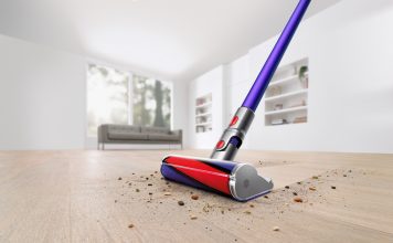 The Dyson V11 Absolute Pro Cordless Stick Vacuum is shown cleaning a hardwood floor in a white room. It makes a clean sweep through the dust and debris on the floor, leaving nothing behind it.