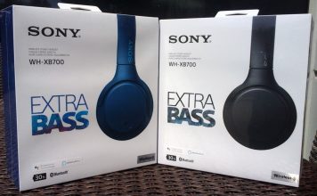 Two white boxes containing Sony WH-XB700 headphones. Each is labeled "SONY WH-XB700 EXTRA BASS." The boxes are outdoors in the sunlight. The left box contains a pair of headphones in blue while the box on the right contains a pair of headphones in black.