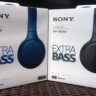 Two white boxes containing Sony WH-XB700 headphones. Each is labeled "SONY WH-XB700 EXTRA BASS." The boxes are outdoors in the sunlight. The left box contains a pair of headphones in blue while the box on the right contains a pair of headphones in black.