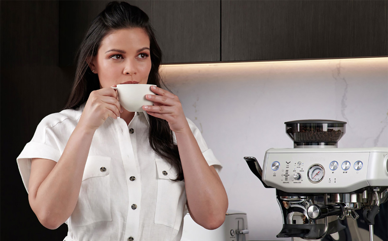 Une femme boit du café avec le Barista Express Impress de Breville en arrière-plan.