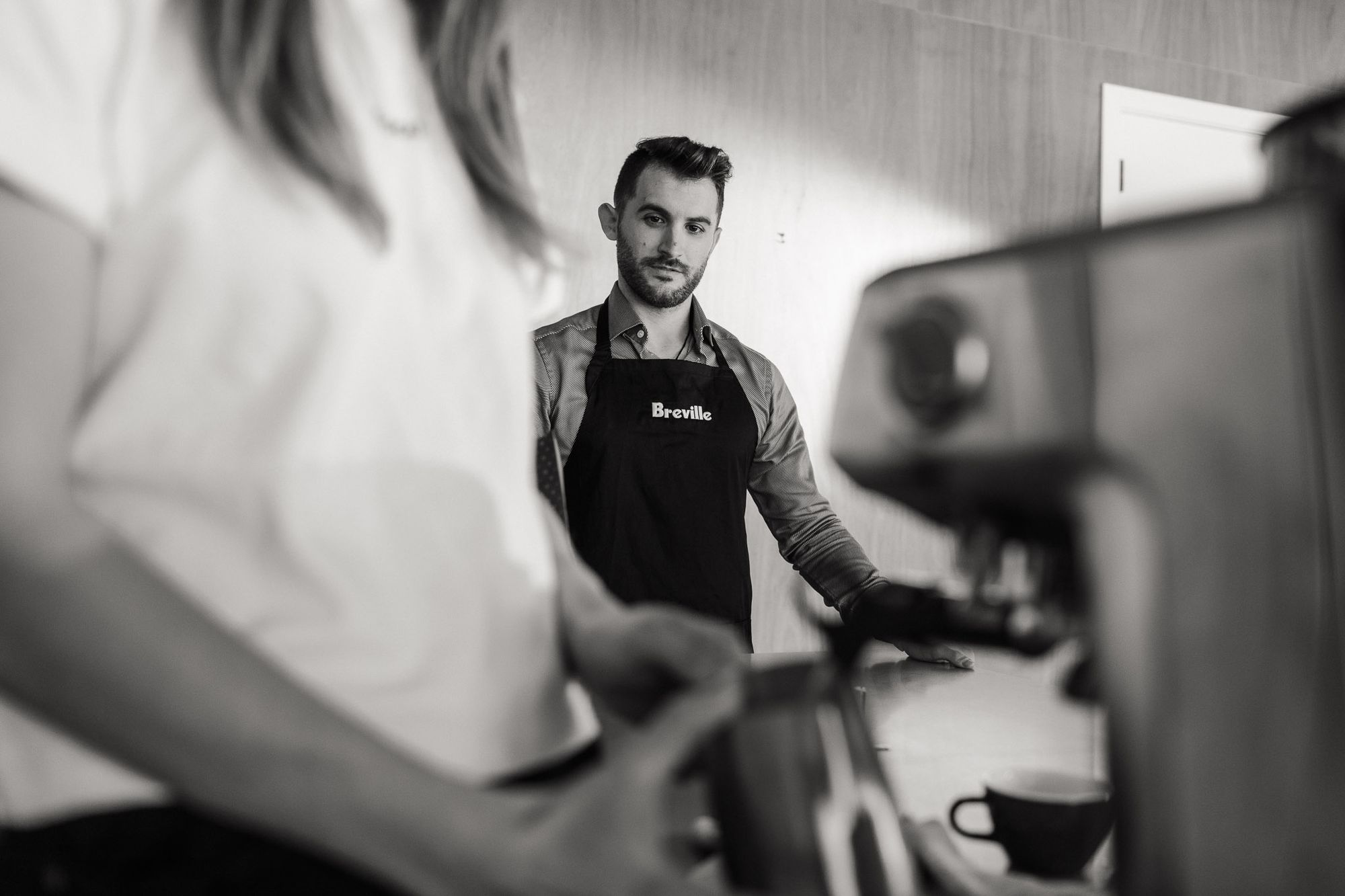 Черно-белое фото: мужчина в фартуке Breville и женщина, готовящая эспрессо из кофемашины Breville.