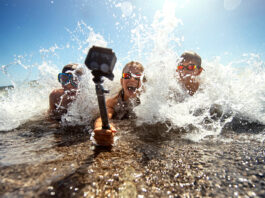 Three young people in swimwear capture a moment of fun with a handheld camera, illustrating tech essentials for an amazing summer as waves splash around them on a sunny beach day.