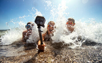 Three young people in swimwear capture a moment of fun with a handheld camera, illustrating tech essentials for an amazing summer as waves splash around them on a sunny beach day.