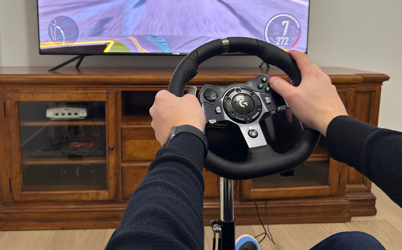 Logitech G920 racing wheel in black with a TV stand and a flat panel TV with reviewer Matthew Rondina's arms reached out and hands on the wheel racing in Forza Motorsport.