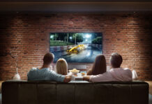 Two couples are cheering while watching game at home. They are sitting on a sofa in the modern living room faced to a big TV set on the front wall. The car is artificial and made in 3D.