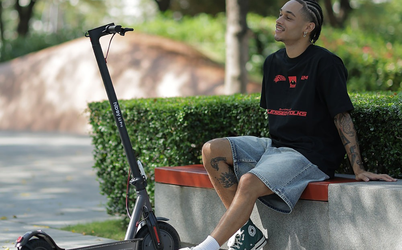 A man sitting by trees with his Hiboy electric scooter in front of him.