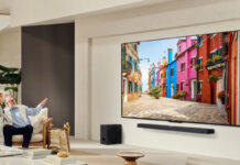 An elderly man with a young girl in a living room pointing at a large LG QNED TV on the wall.