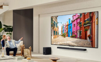 An elderly man with a young girl in a living room pointing at a large LG QNED TV on the wall.