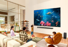 A family sitting around a living room watching an aquarium scene on a large LG QNED TV on the wall.