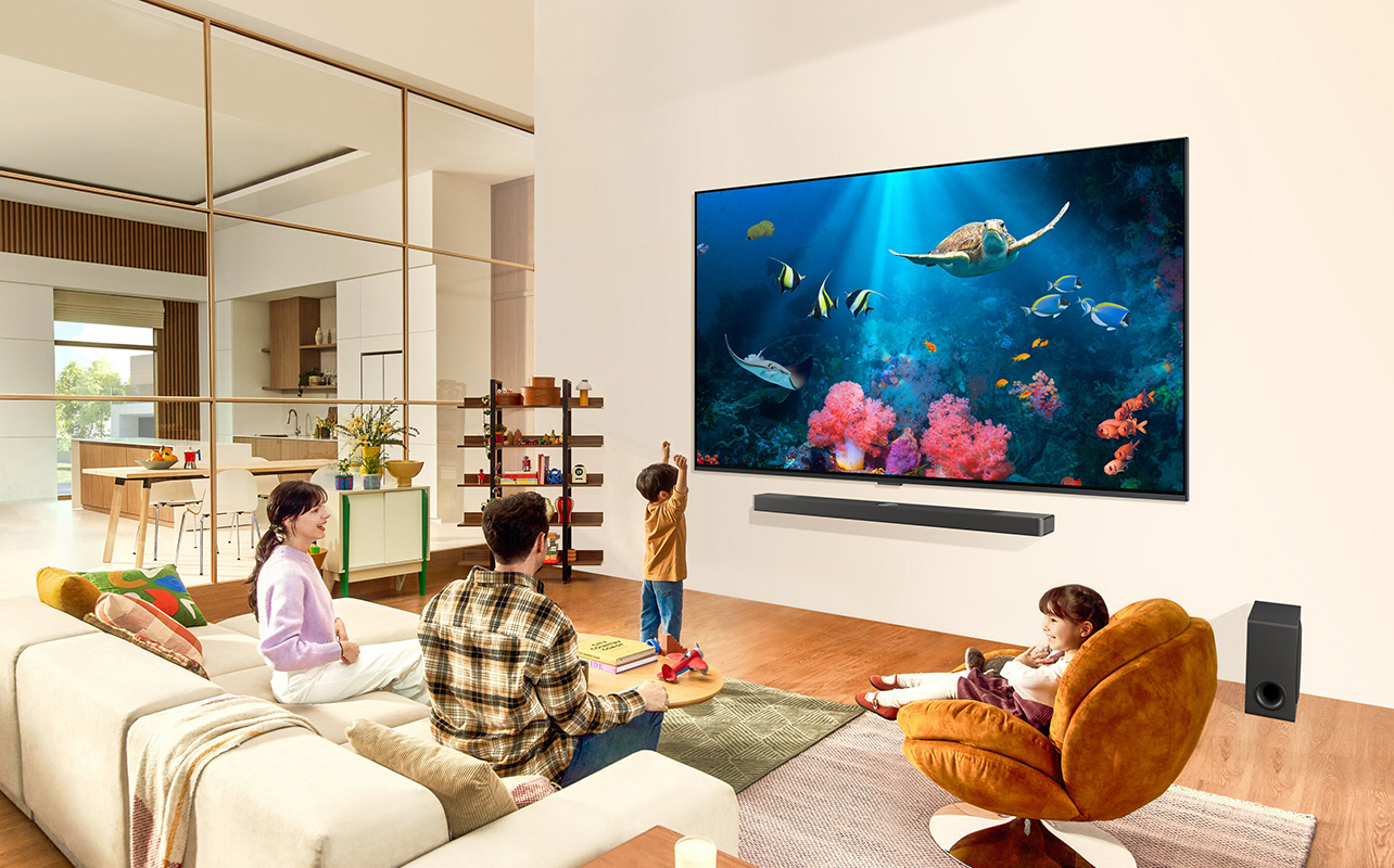 A family sitting around a living room watching an aquarium scene on a large LG QNED TV on the wall.