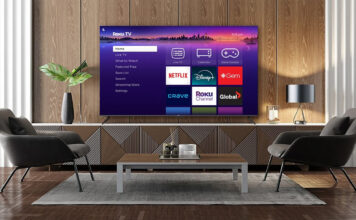 A Roku TV between two chairs and a table, showing the smart TV home interface.