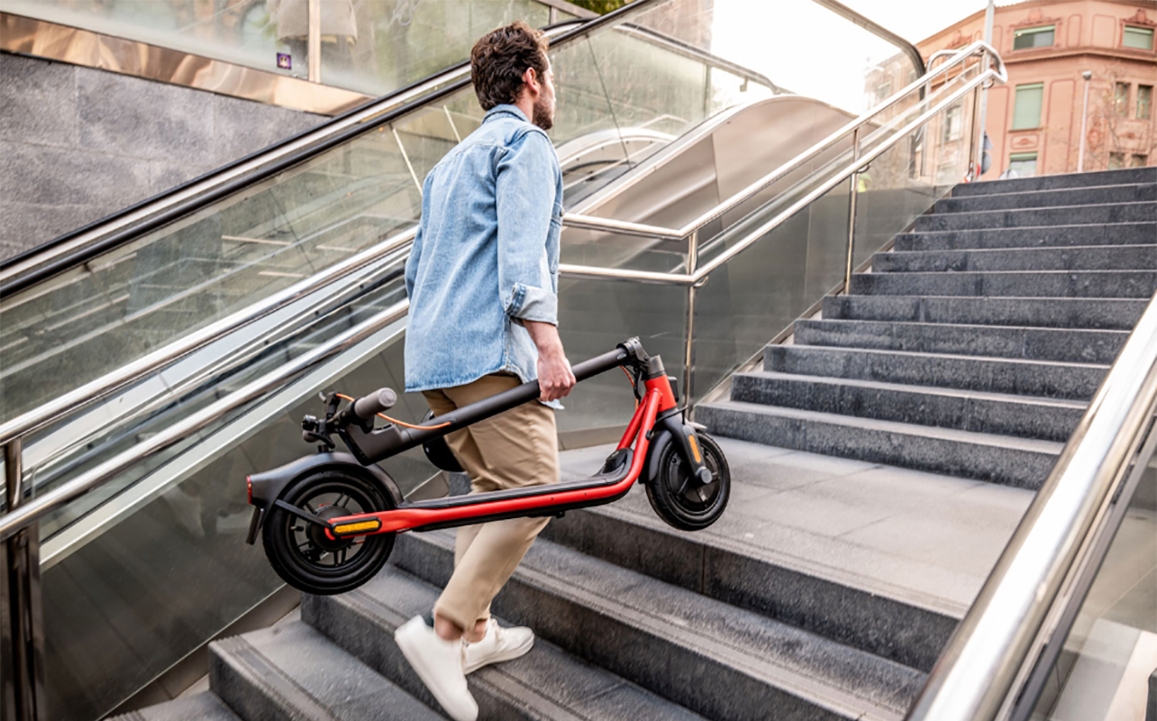 A man carrying a folded up Segway Ninebot e-scooter up a flight of outdoor steps.