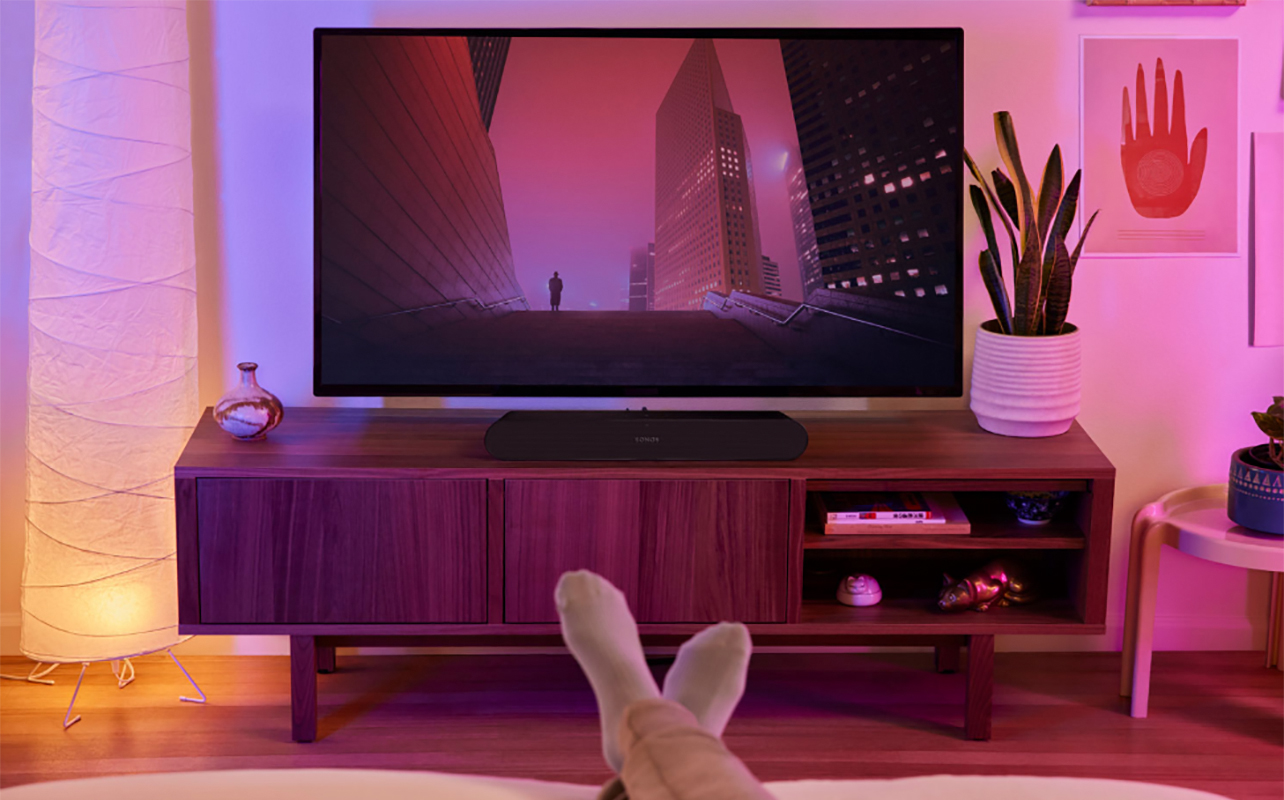 A Sonos Ray sound bar beneath a TV with feet relaxing on the table in view in front of it.