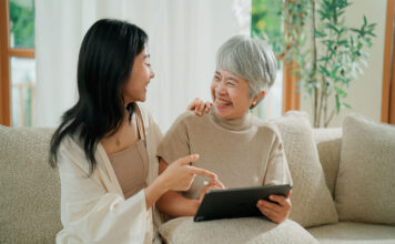 A young Asian woman teaching her retired mother how to use a digital tablet in the living room.
