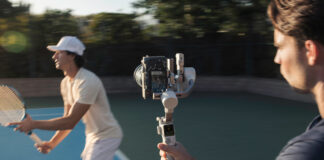 Man using camera on gimbal to track tennis player.