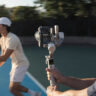 Man using camera on gimbal to track tennis player.