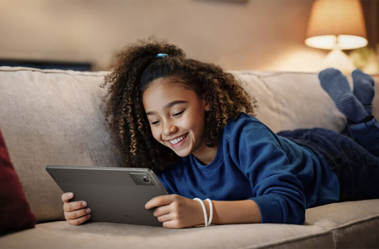 A young girl lying on the couch smiling while holding a Lenovo Tab M11 tablet.