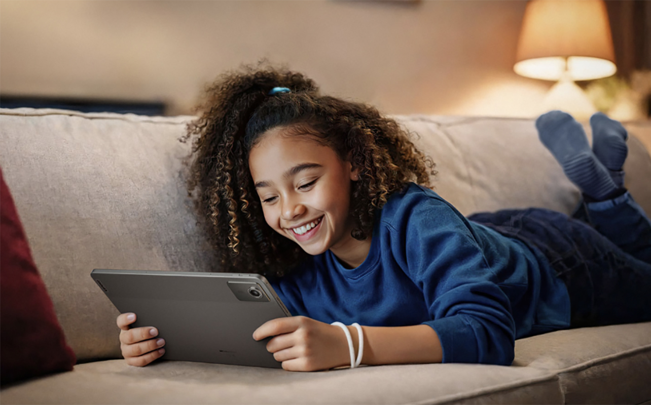 A young girl lying on the couch smiling while holding a Lenovo Tab M11 tablet.