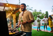 Man grilling food while holding a beer at an outdoor barbecue party by the pool. Group of friends socializing in the background. Summer celebration concept. Medium shot