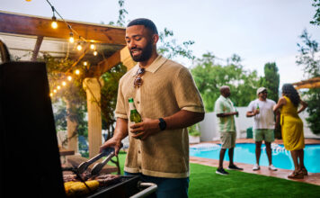 Man grilling food while holding a beer at an outdoor barbecue party by the pool. Group of friends socializing in the background. Summer celebration concept. Medium shot
