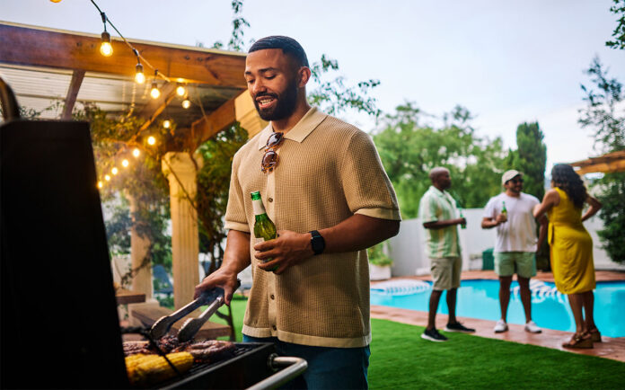Man grilling food while holding a beer at an outdoor barbecue party by the pool. Group of friends socializing in the background. Summer celebration concept. Medium shot
