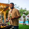 Man grilling food while holding a beer at an outdoor barbecue party by the pool. Group of friends socializing in the background. Summer celebration concept. Medium shot