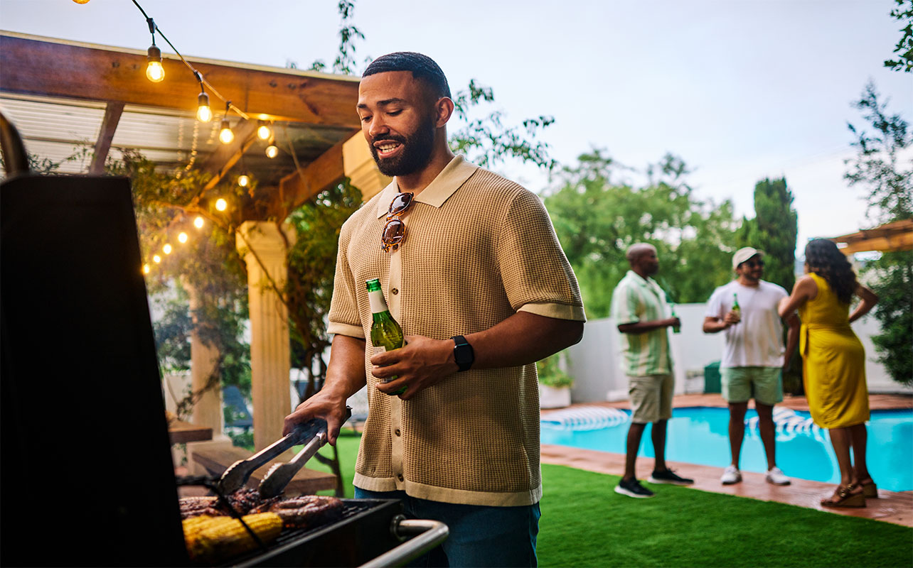 Man grilling food while holding a beer at an outdoor barbecue party by the pool. Group of friends socializing in the background. Summer celebration concept. Medium shot
