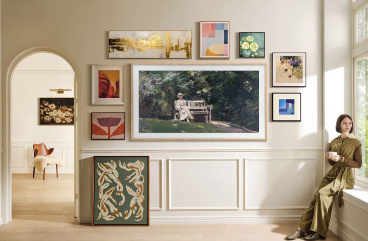 A Samsung The Frame TV mounted on the wall with smaller picture frames around it, a woman sitting on the windowsill to the right.