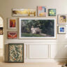 A Samsung The Frame TV mounted on the wall with smaller picture frames around it, a woman sitting on the windowsill to the right.