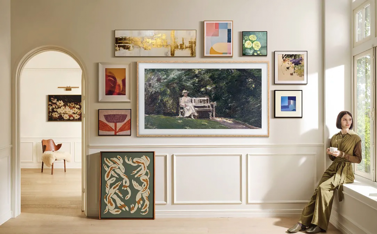 A Samsung The Frame TV mounted on the wall with smaller picture frames around it, a woman sitting on the windowsill to the right.