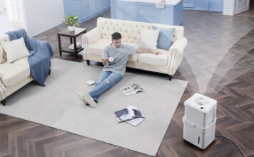 An overhead view of a man sitting on a living room floor with a Midea Cube dehumidifier in the bottom, right corner.