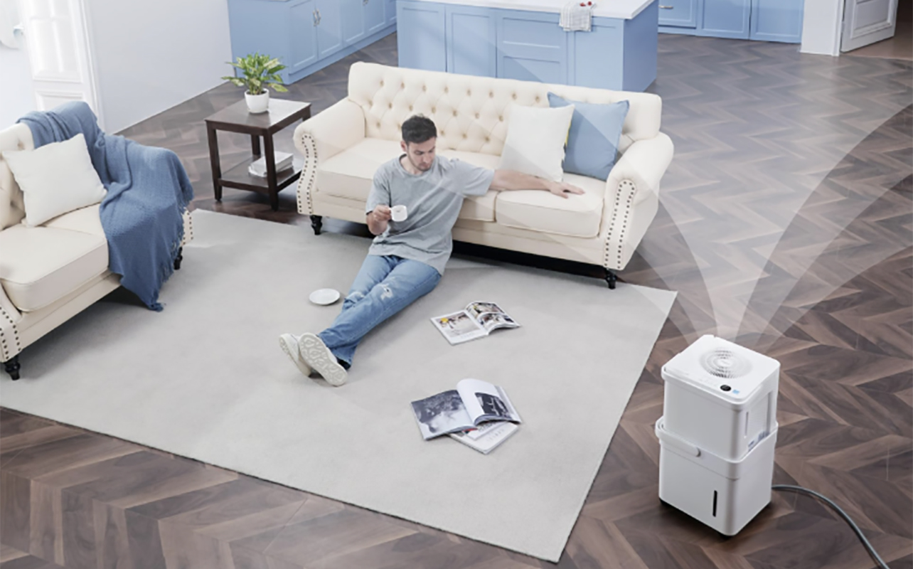 An overhead view of a man sitting on a living room floor with a Midea Cube dehumidifier in the bottom, right corner.