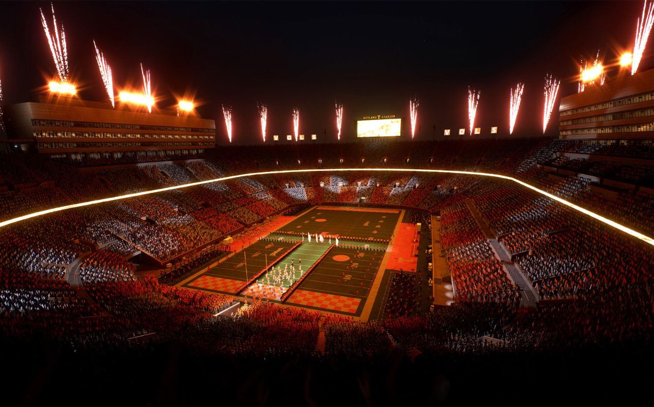 Fireworks illuminate a packed stadium at night, with crowds, a lit field, and "VOLS" text on display. Festive and energetic atmosphere.
