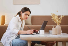 Woman taking notes on a pad next to a laptop on living room table.