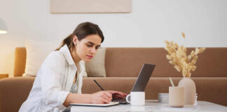 Woman taking notes on a pad next to a laptop on living room table.
