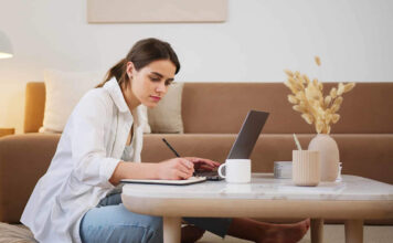 Woman taking notes on a pad next to a laptop on living room table.