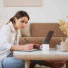 Woman taking notes on a pad next to a laptop on living room table.