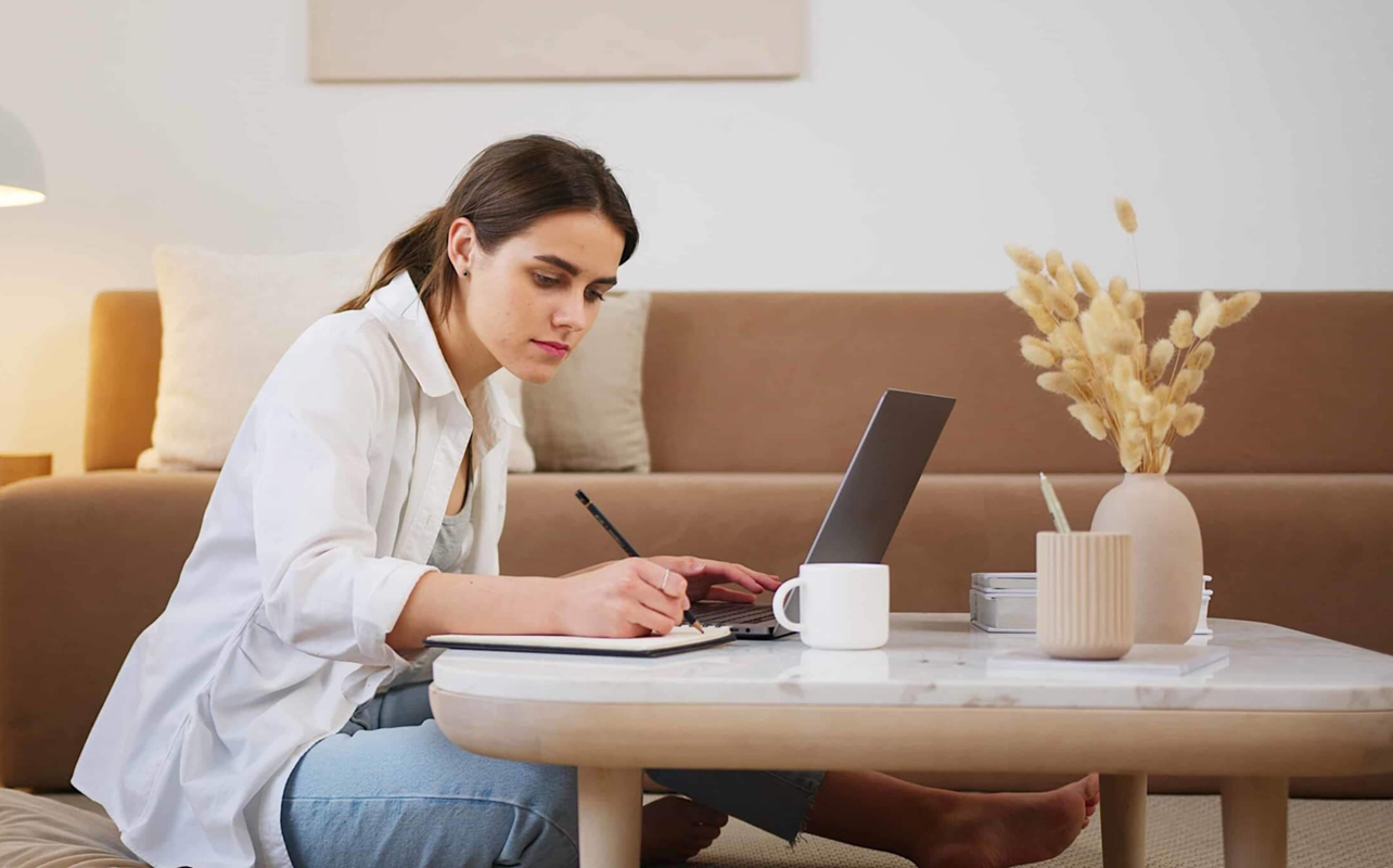 Woman taking notes on a pad next to a laptop on living room table.