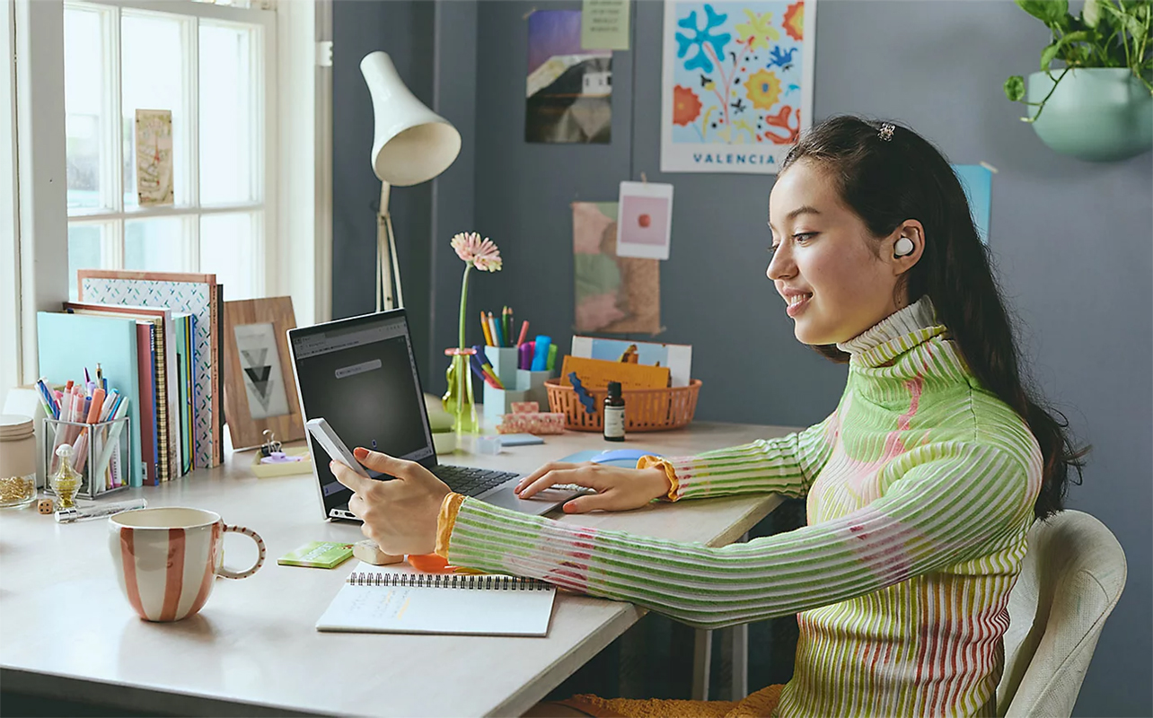 Young student wearing Sony earbuds in front of laptop with phone in hand at home.