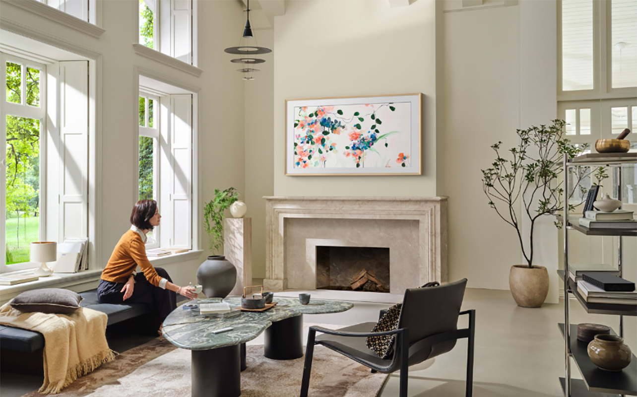 a woman sitting by a window looking at a Samsung The Frame TV mounted above a fireplace, showing artwork.
