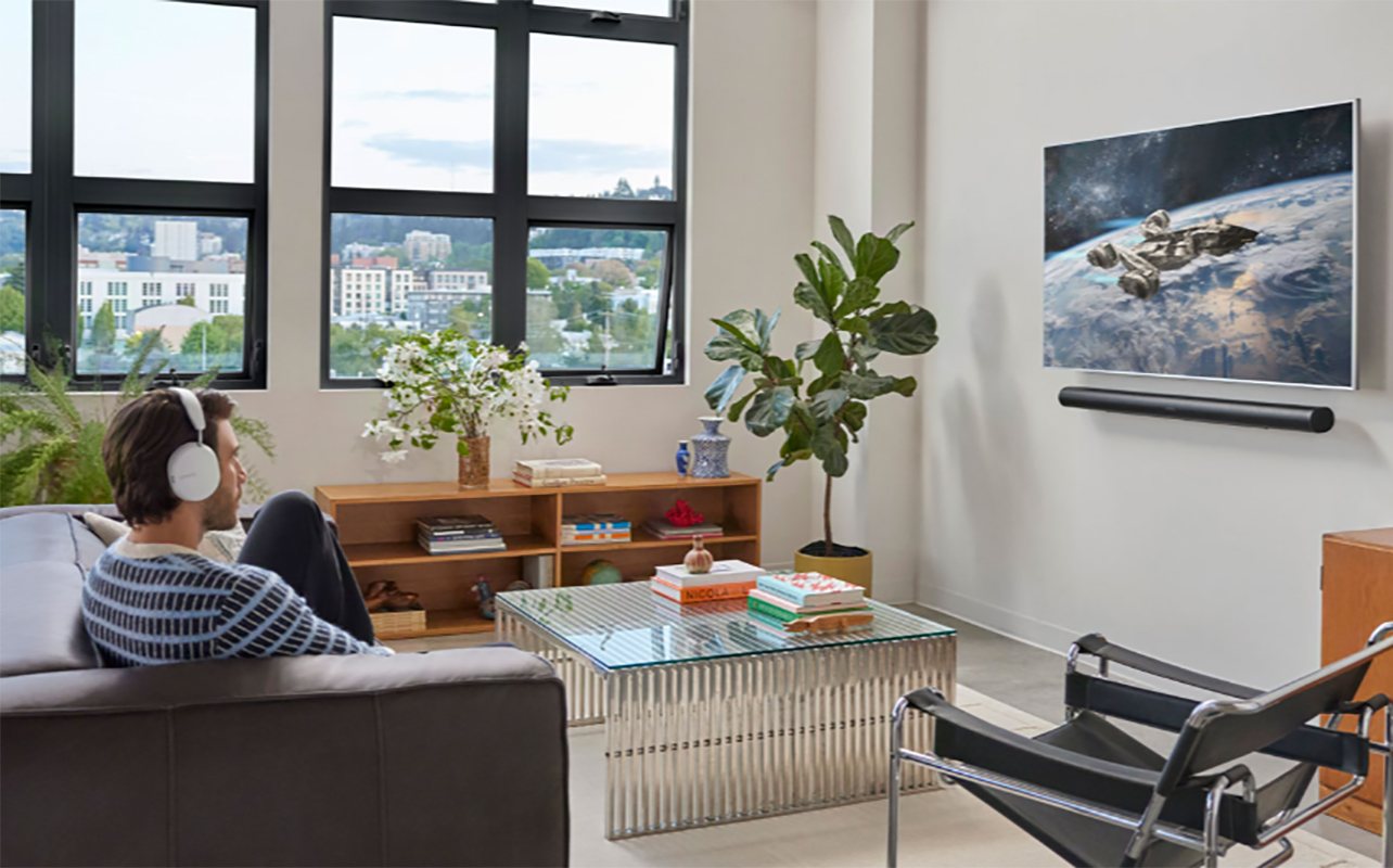 A man sitting down watching TV wearing Sonos Ace headphones with the Sonos Arc Ultra soundbar mounted under the TV.
