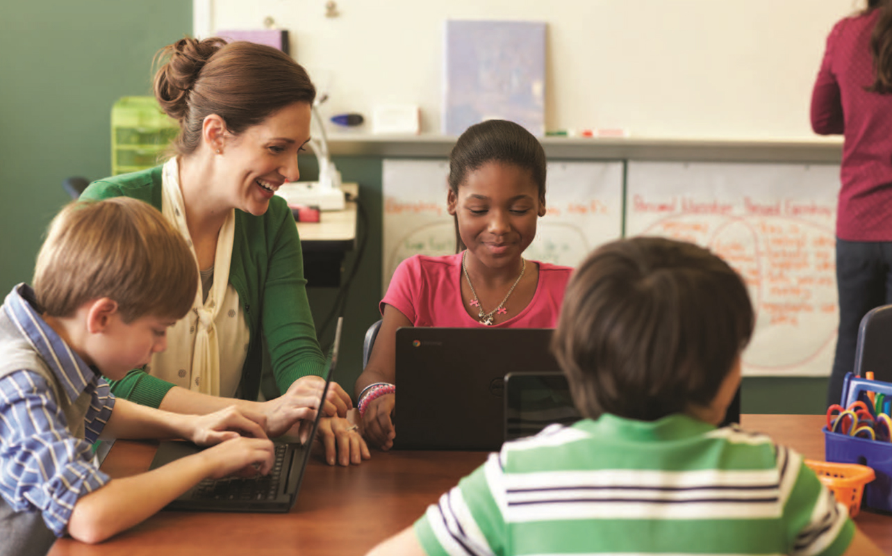Young students in a classroom using Chromebooks with a teacher looking on.