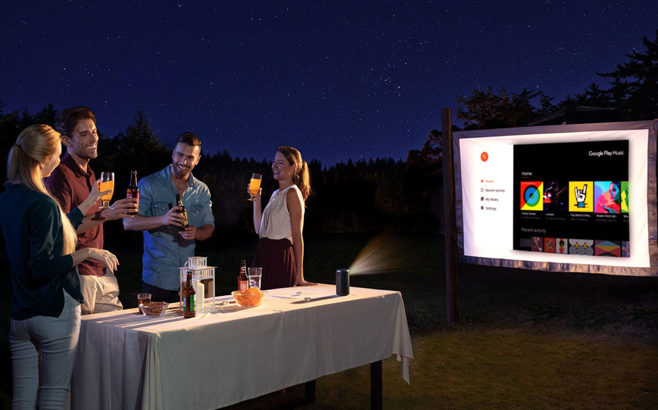People by a table outside talking with an Anker Nebula Capsule II projector displaying an image on an outdoor screen.