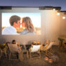 Four people sitting outside on folding chairs in front of a screen using an Xgimi portable projector.