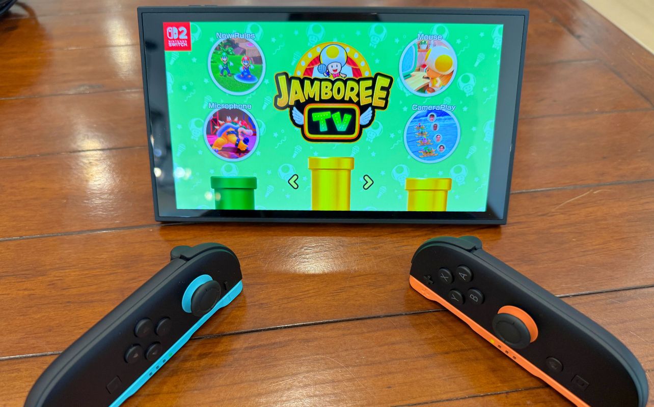Nintendo Switch 2 screen shows "Jamboree TV" game on a green background. Two controllers lay on a wooden surface, one blue, one orange.